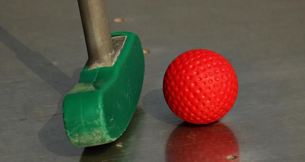 Green putter head next to a red golf ball on a silver surface.