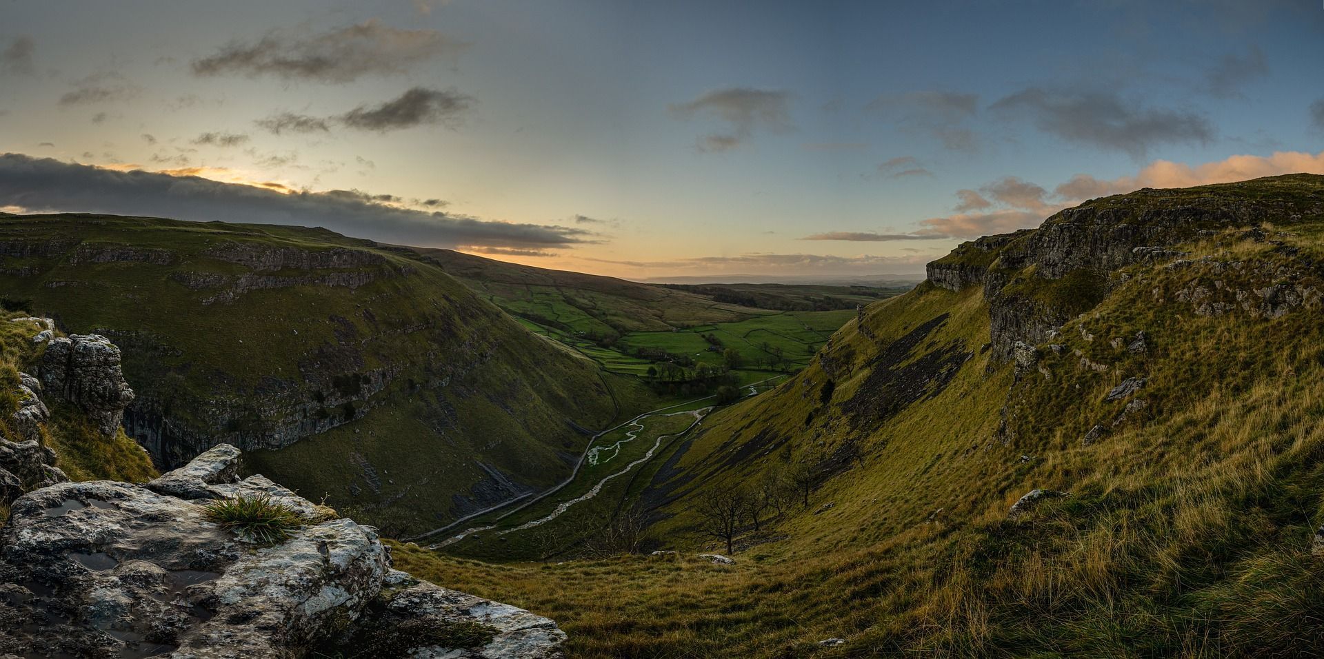 Valley landscape with grassy hills under a cloudy sky at sunset.
