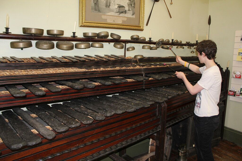 Person playing a large antique musical instrument with bars and bells in a room.
