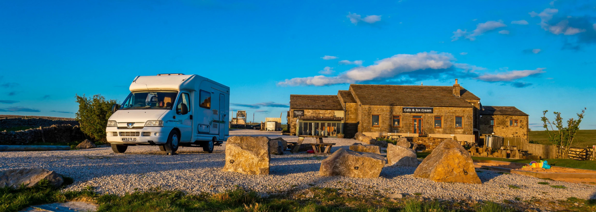 A white camper van on a gravel road, passing a stone building under a blue sky.