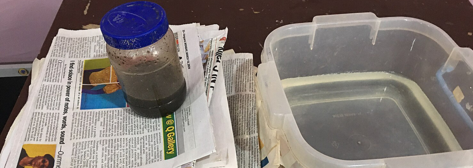 A jar with a blue lid sits on newspaper next to a clear plastic container with water.