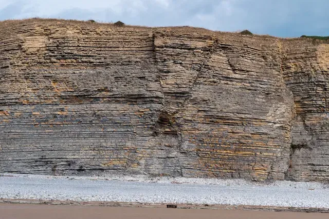 Layered sandstone cliff face with light and dark bands, overlooking a pebble beach.