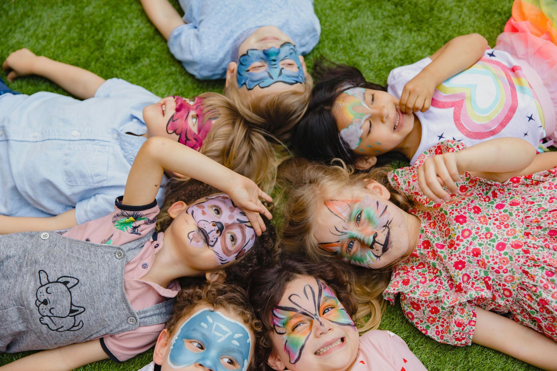 Children with face paint lying on grass, heads together.