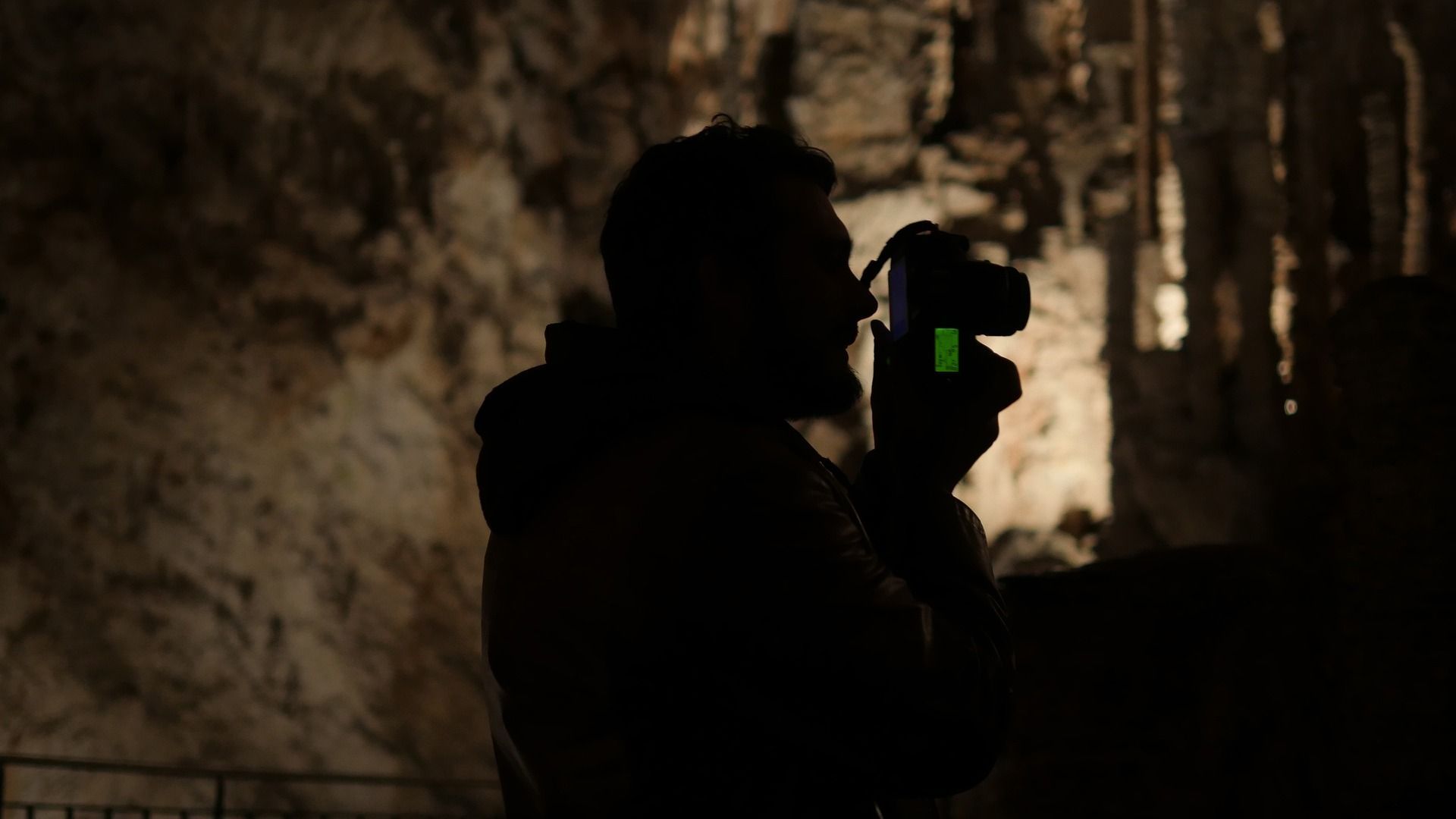 Silhouette of a person holding a camera in a dark cave, illuminated by a camera's green light.