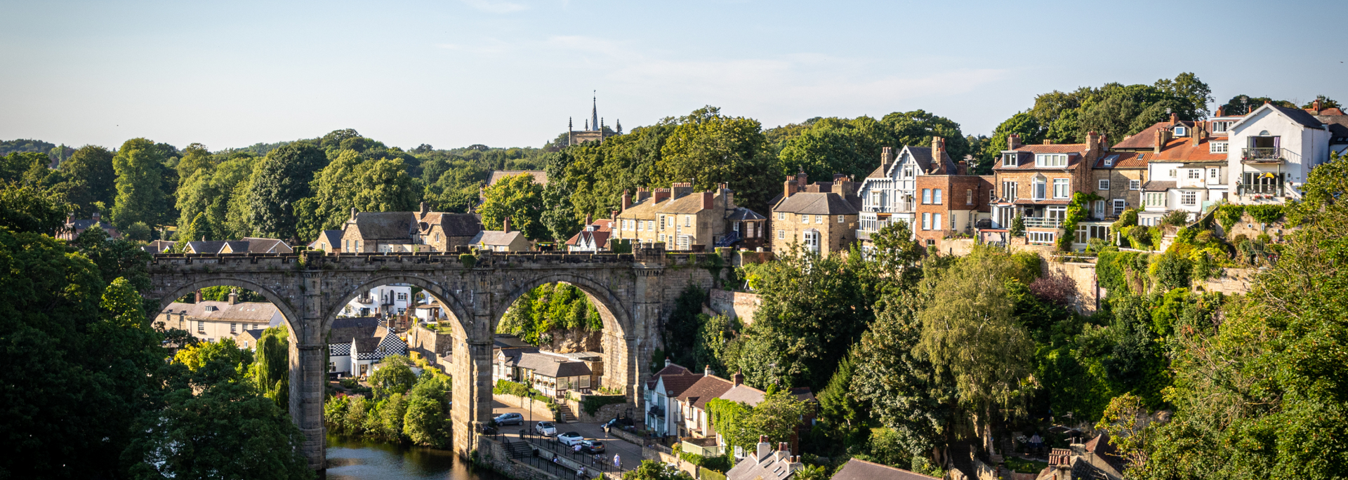Stone bridge arches over river in a lush green valley with houses on the hills.