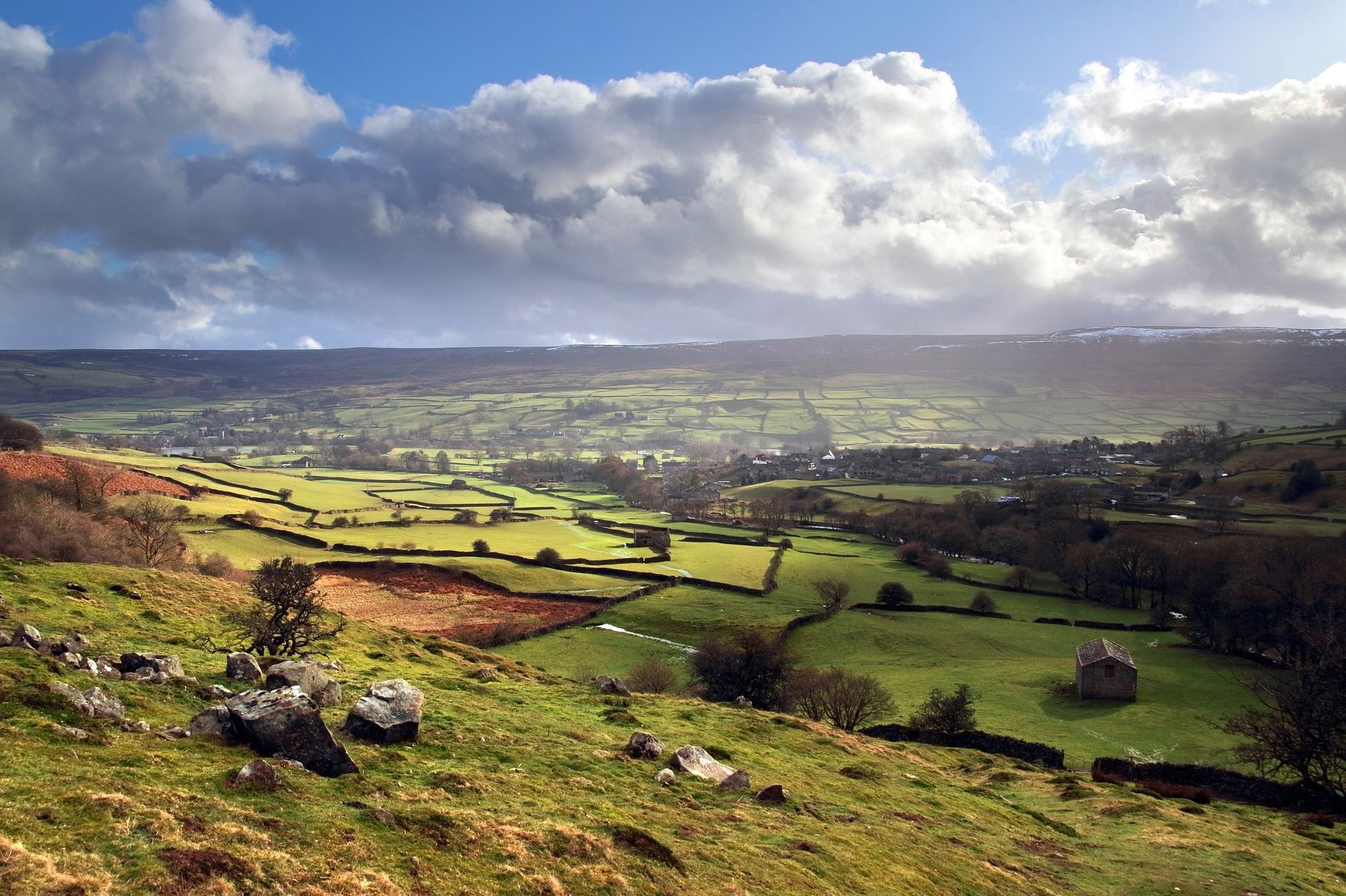 Rolling green valley under cloudy sky. Farmland, stone walls, and trees. Sunlight breaking through.