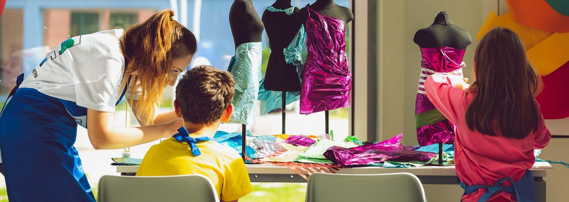 A woman and two children working on dressmaking projects. Dresses on mannequins are visible in the background.