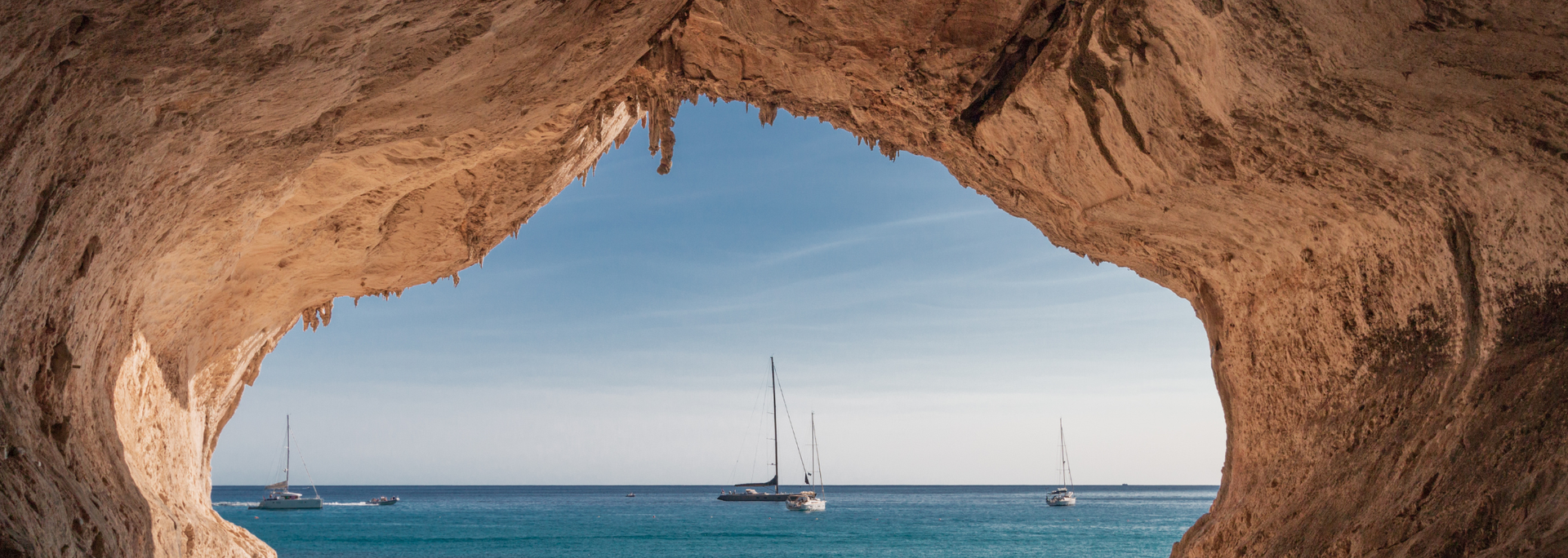 View of the ocean and sailboats framed by a natural rock arch. Blue water and sky.