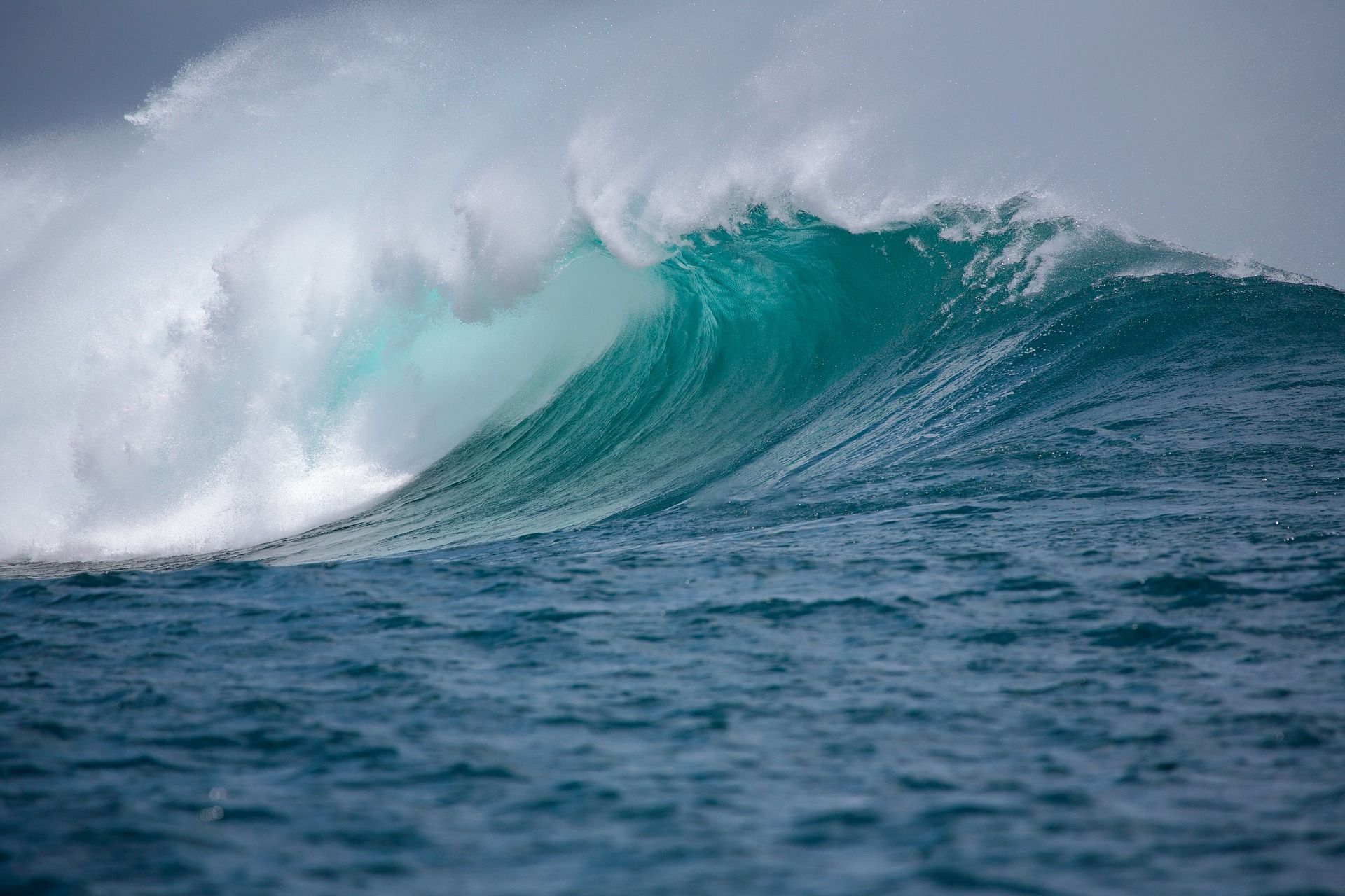 A large, turquoise ocean wave crests and curls over, white foam against a cloudy sky.
