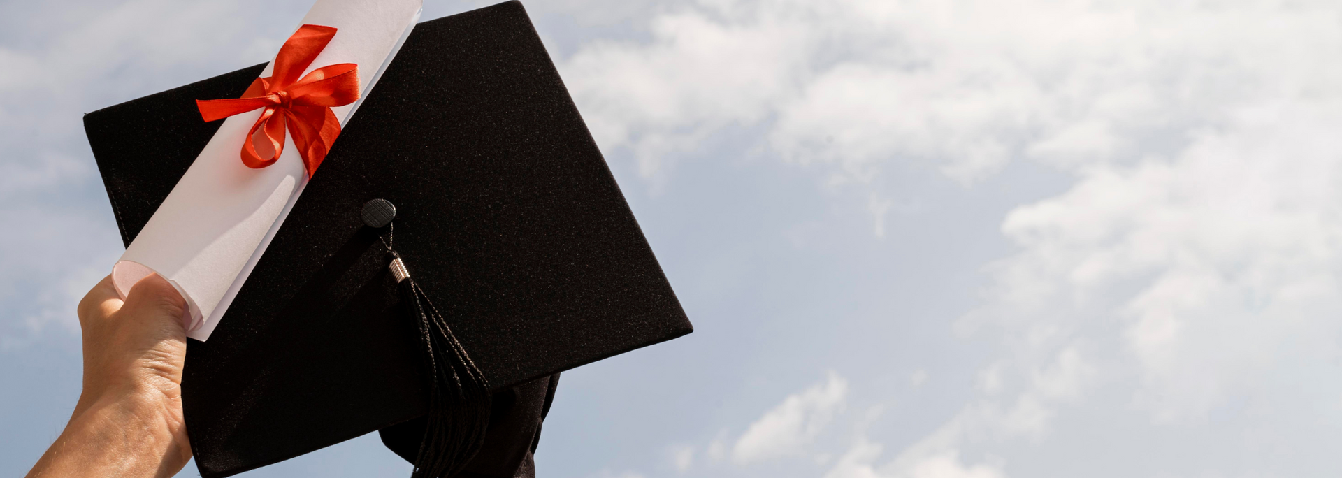 Hand holding diploma tied with a red ribbon, with a graduation cap against a cloudy sky.