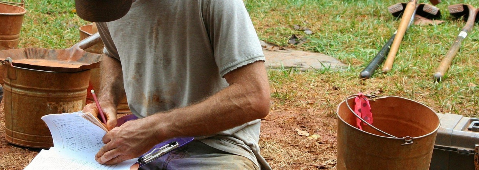 Person writing on paper, near buckets and tools in an outdoor setting.