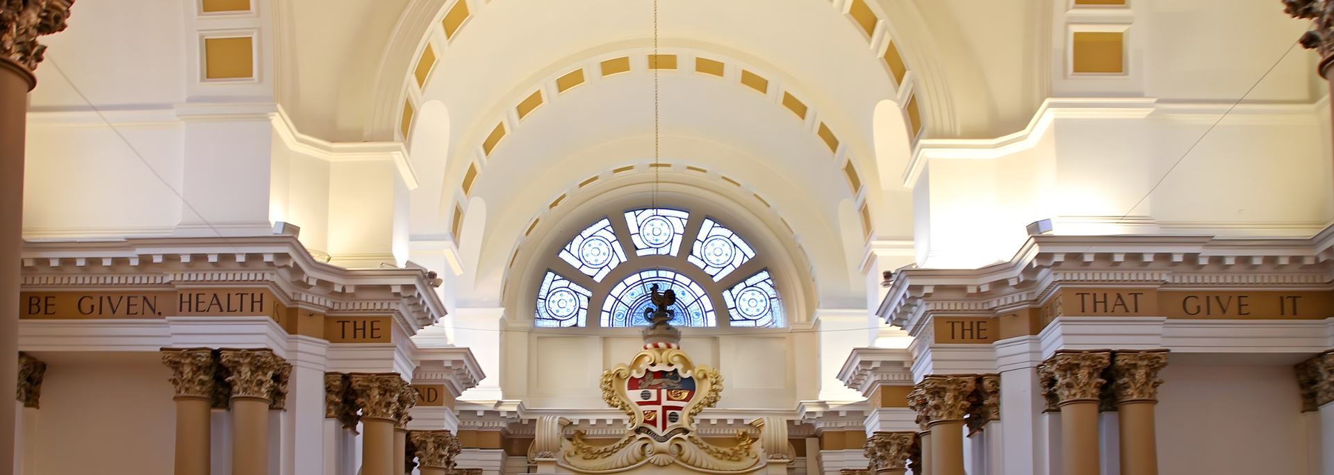 Interior of a cathedral with a stained-glass window above a crest, columns, and arched ceiling.