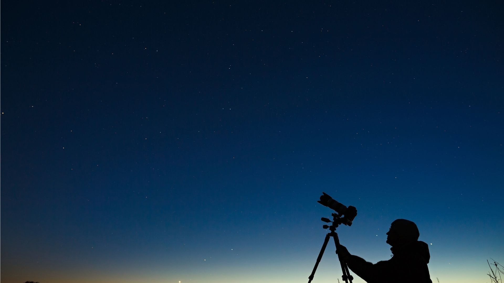 Silhouette of a person using a telescope under a dark blue sky with visible stars.