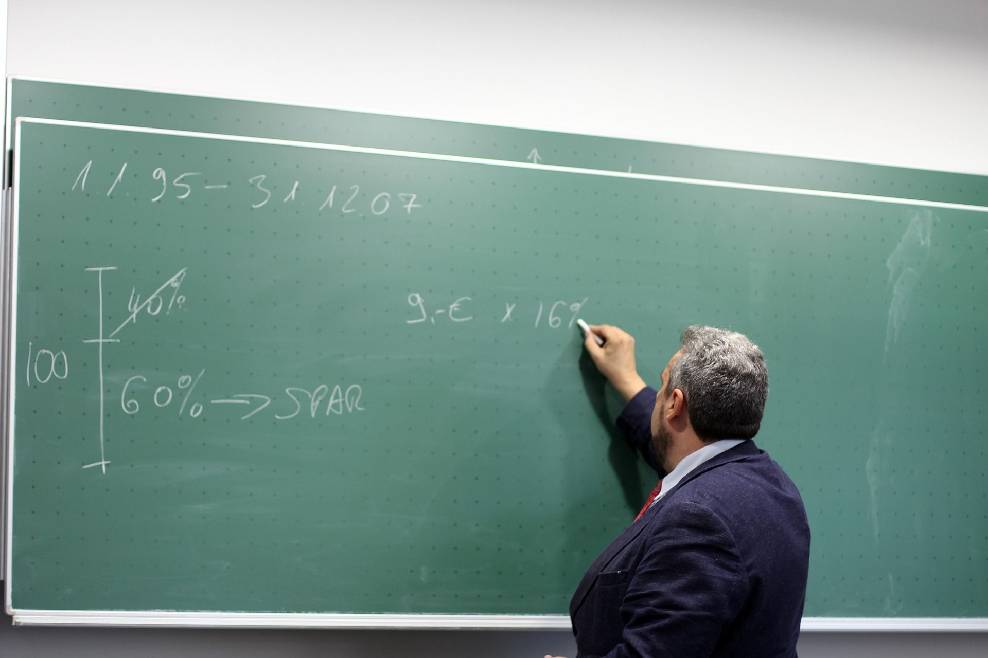 Man writing math equations on a green chalkboard; classroom setting.