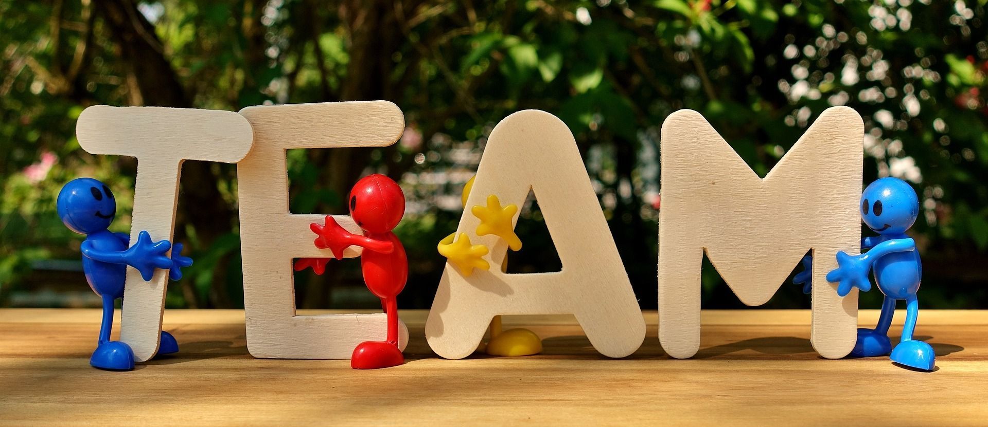 Three figures supporting the word TEAM. The blue figures hold the letters T and M. Red figure holds the letter E. The letter A has yellow embellishments.