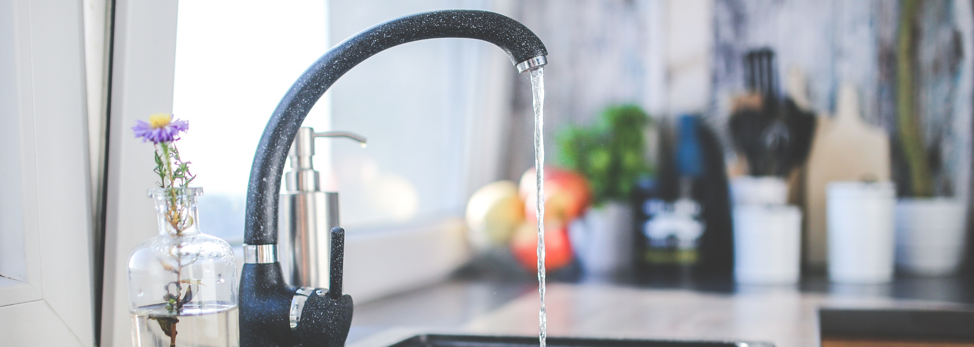 Kitchen faucet with running water; vase with purple flower on countertop.