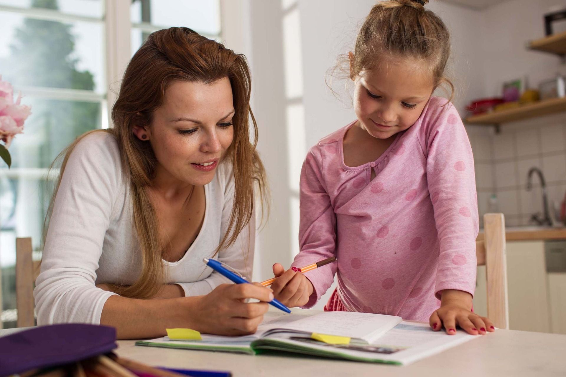Woman and young girl at a table, writing in a notebook. Woman holds a blue pen, child holds a pencil.
