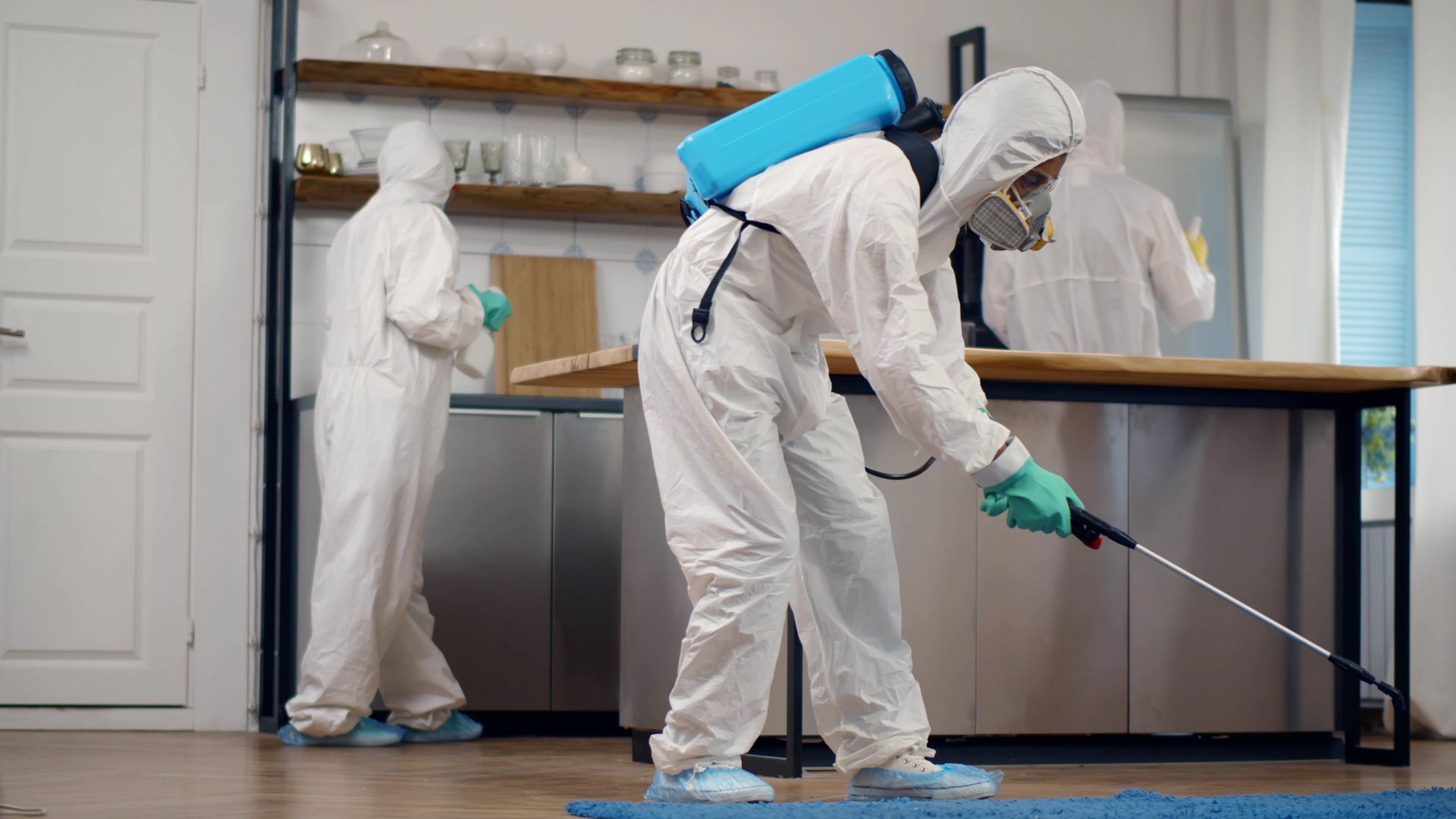 Two people in hazmat suits disinfecting a kitchen. One sprays the floor, another wipes a counter.