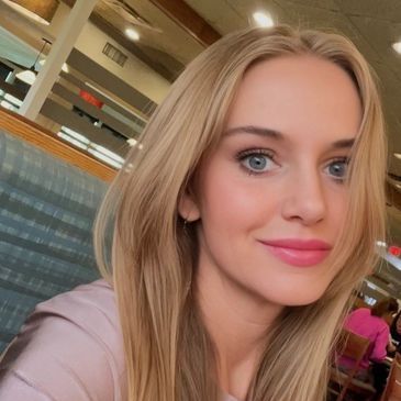 Blonde woman with blue eyes smiles. Indoors, she wears pink lipstick and a light purple shirt.