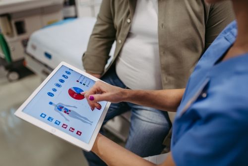 Nurse pointing at a digital tablet with medical diagrams; patient seated nearby.