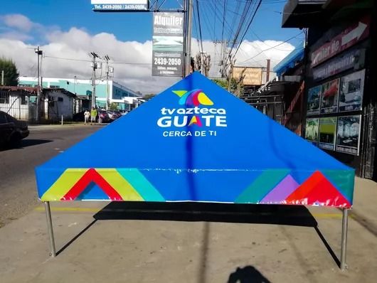 Carpa azul para eventos con el logotipo de “tv azteca GUATE” y franjas coloridas en una calle bajo un cielo despejado.