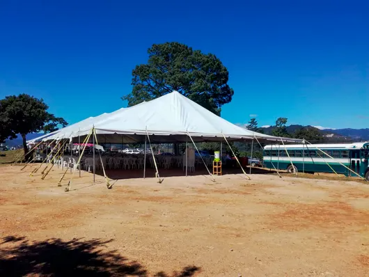 Carpa blanca para eventos en un campo polvoriento bajo un cielo azul brillante, con árboles y montañas al fondo.