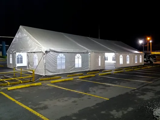 Una gran carpa blanca instalada de noche en un estacionamiento con luces brillantes y barandillas amarillas cerca.
