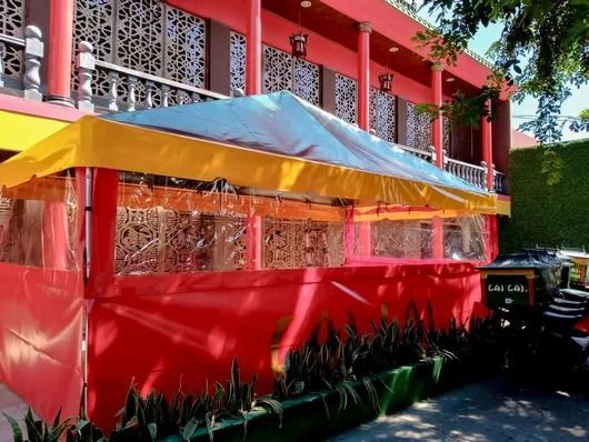 Edificio rojo con toldo amarillo y plantas frondosas en la base; vehículo estacionado a la derecha.