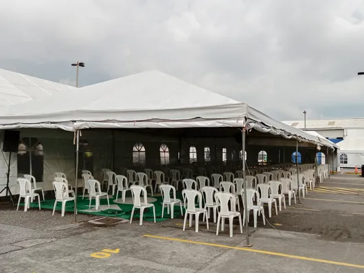 Filas de sillas blancas bajo una gran carpa blanca en un estacionamiento.