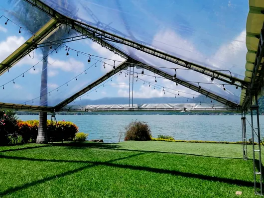 Césped resguardado junto al agua con toldo metálico, agua azul y un puente a lo lejos bajo un cielo despejado.