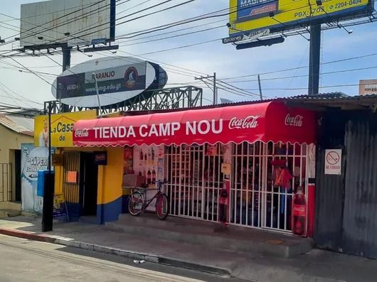 Vista de la fachada de la tienda Tienda Camp Nou con toldo rojo y puerta de seguridad metálica.
