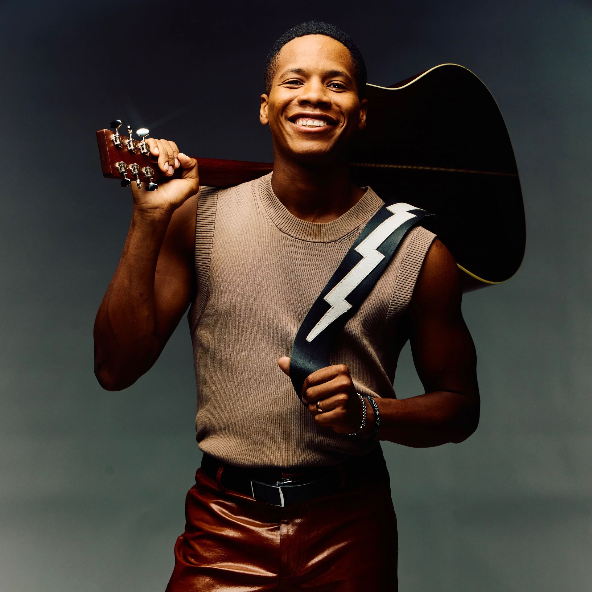 A smiling Thunderstorm Artis wears a beige sleeveless top and leather pants, holding an acoustic guitar over their shoulder.