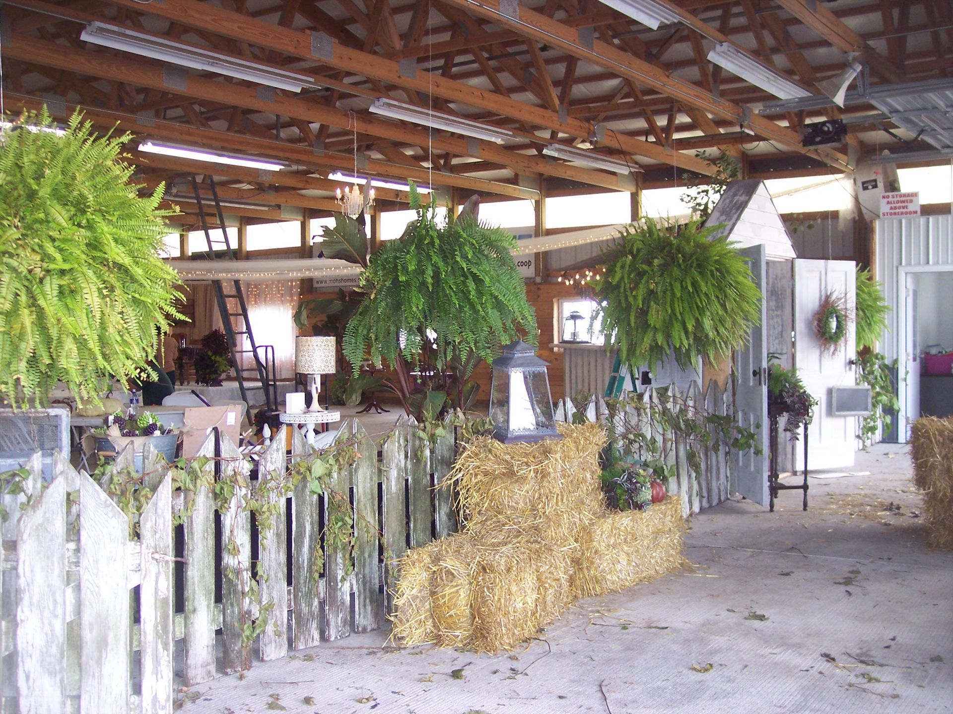 Rustic wedding-style decor in an indoor barn, featuring white picket fences, hay bales, hanging ferns, and lanterns.