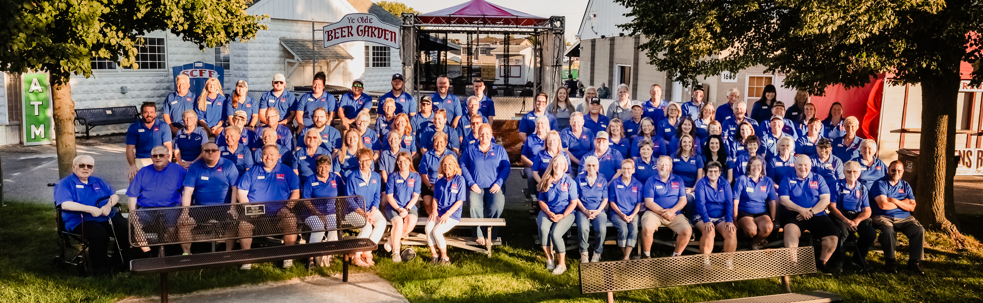 Large group of people in blue shirts posing outdoors near buildings, some seated.