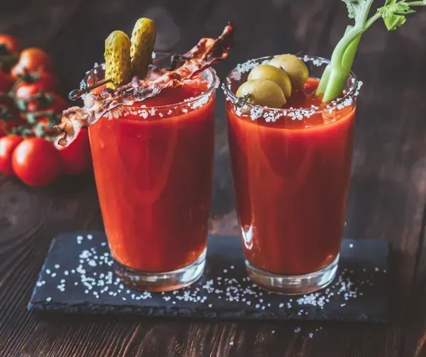 Two Bloody Mary cocktails garnished with bacon, pickles, olives, celery, and salt rims.