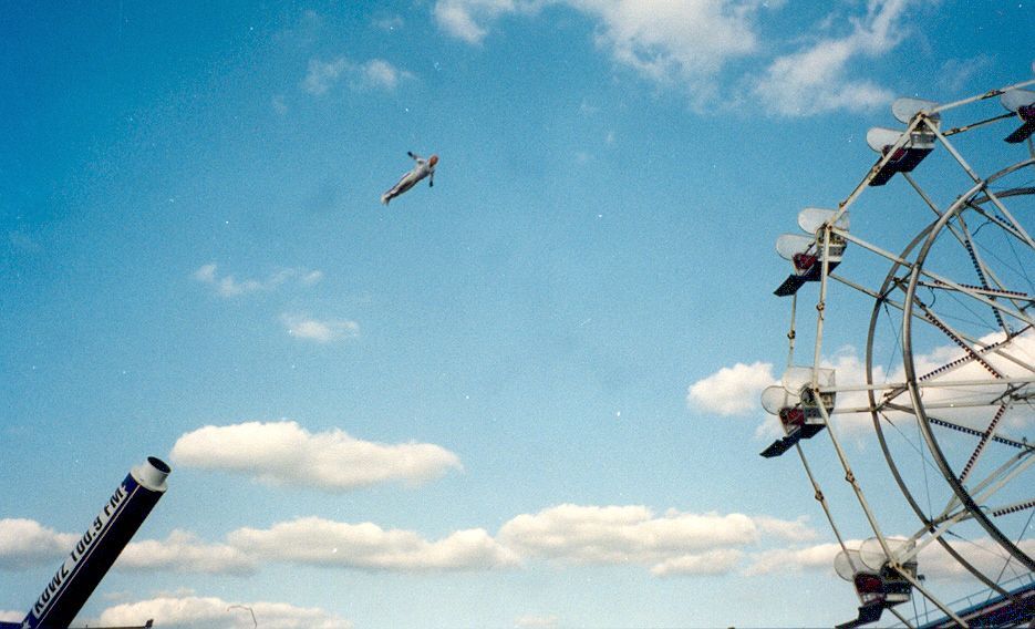 Human cannonball flying over a Ferris wheel on a partly cloudy day.