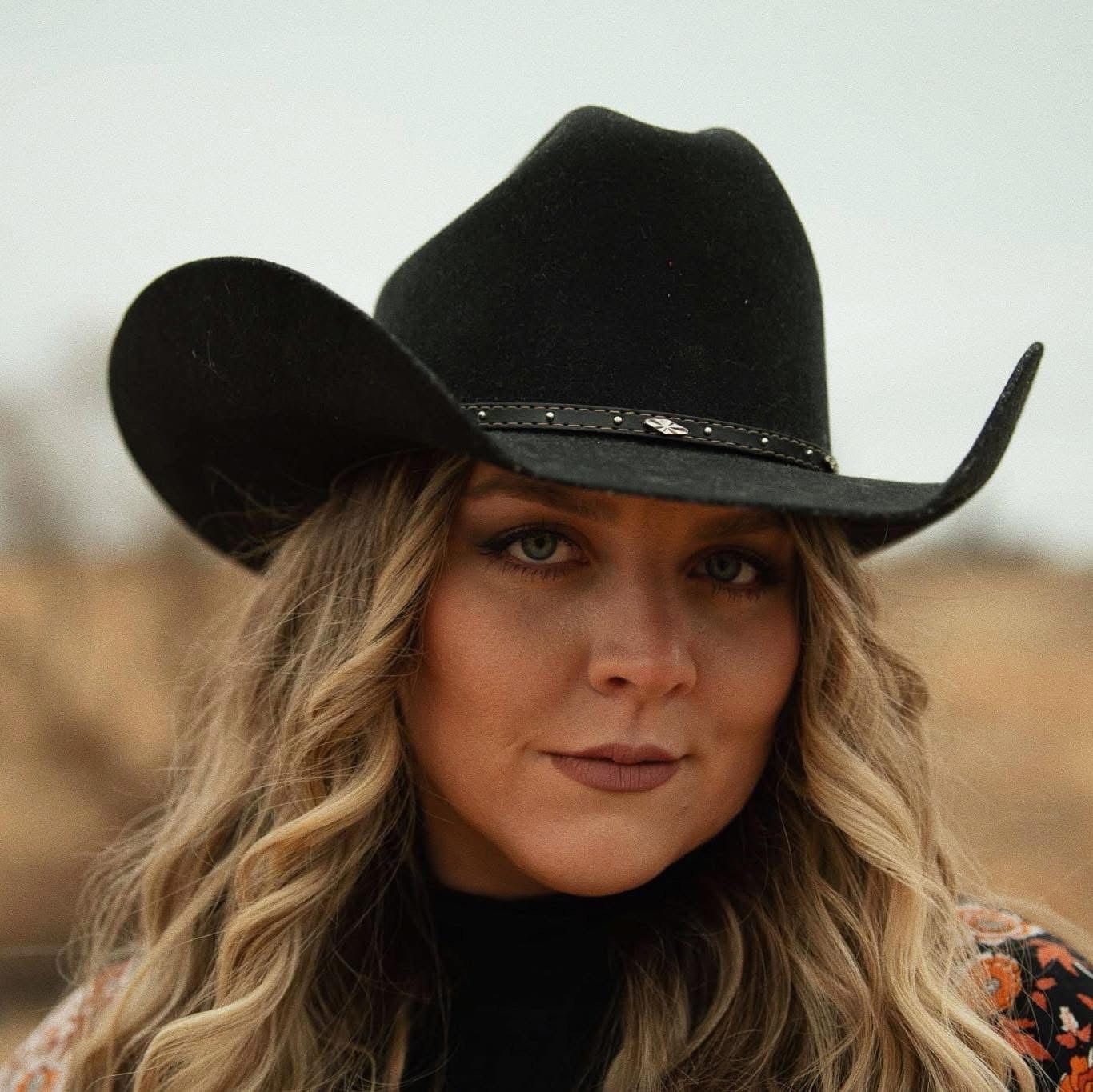 Cori Kennedy in a black cowboy hat with wavy hair, looking at the camera outdoors.