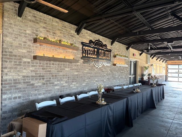 A long table with black tablecloths and white chairs set up in front of a brick wall marked with a