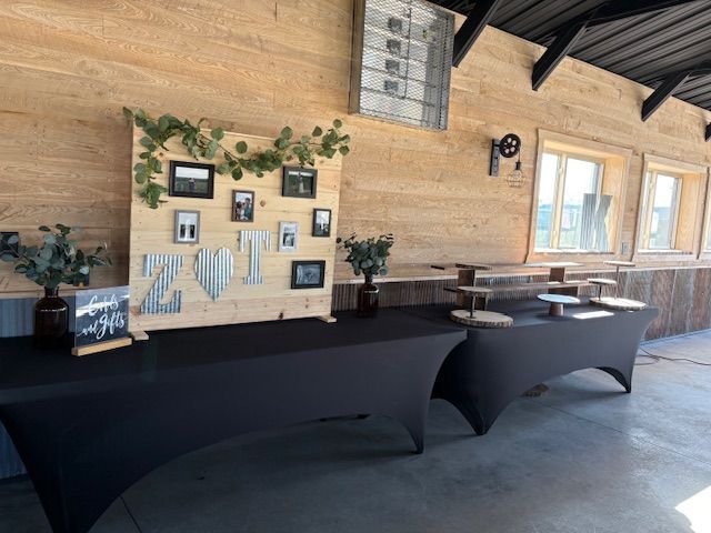 A rustic wood display board with photos and metal letters sits on a black-covered table in a barn-style venue.