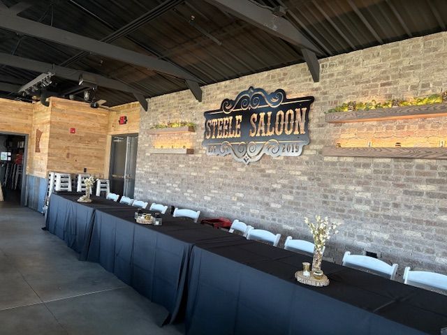 A long table with a black tablecloth and white chairs inside a room with a rustic brick wall and Steele Saloon sign.