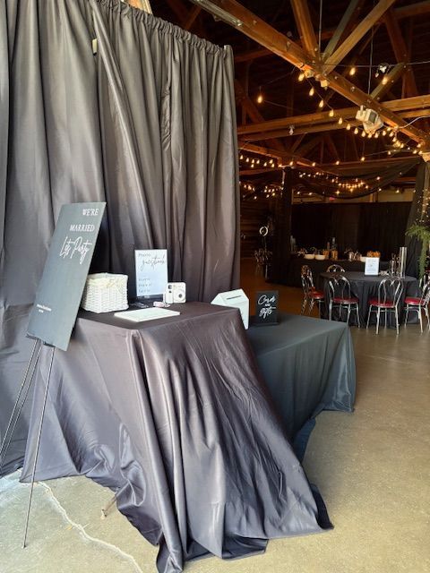 A draped table serves as a wedding guest book station, with a sign on an easel in a rustic venue with string lights.