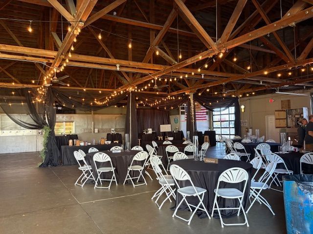 Event space set up with round tables and white chairs under wooden rafters decorated with string lights.