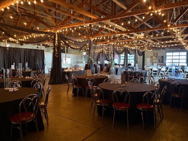 A dimly lit indoor event space featuring round tables with black tablecloths and string lights hanging from wooden rafters.
