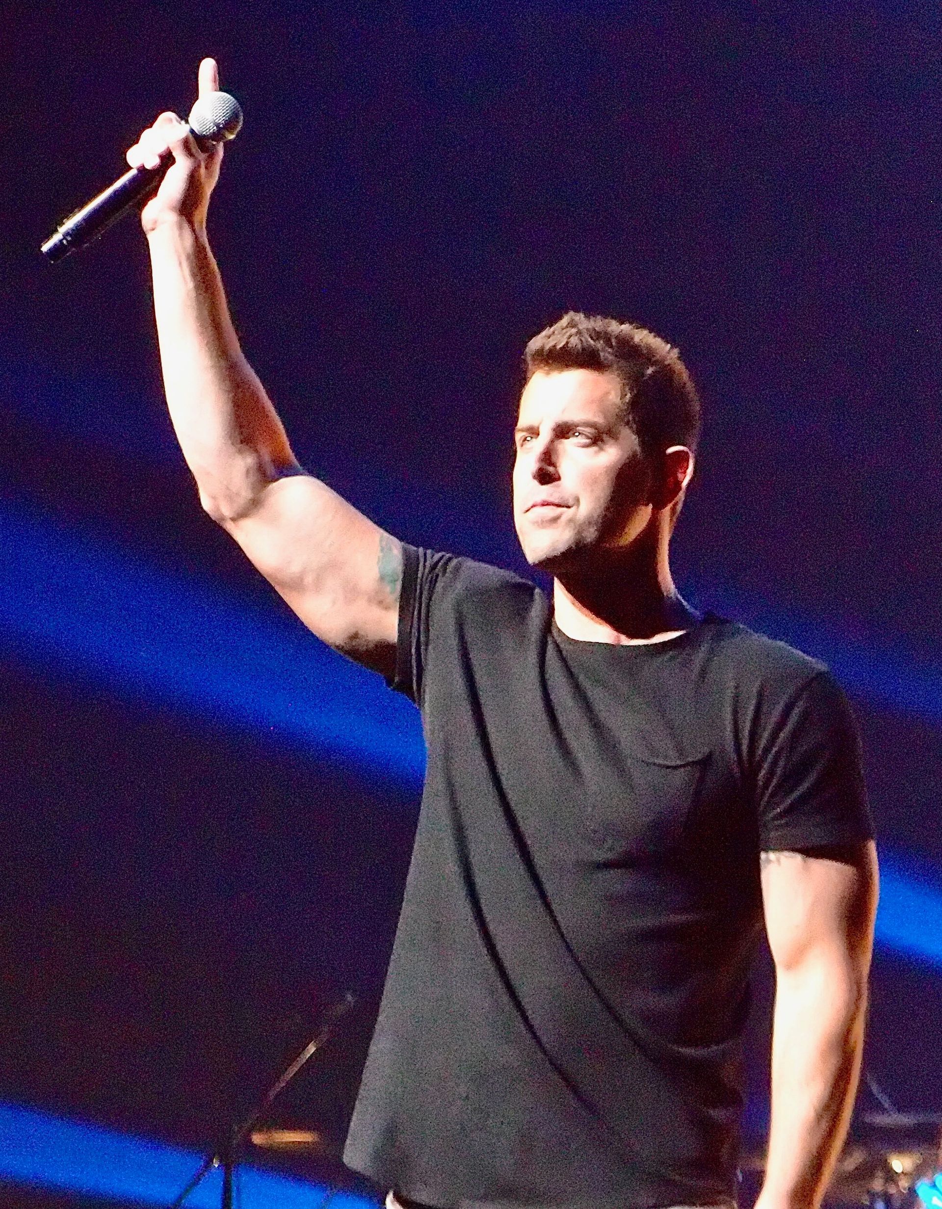 Jeremy Camp in a black t-shirt stands on stage with one arm raised holding a microphone, lit by blue stage lights.