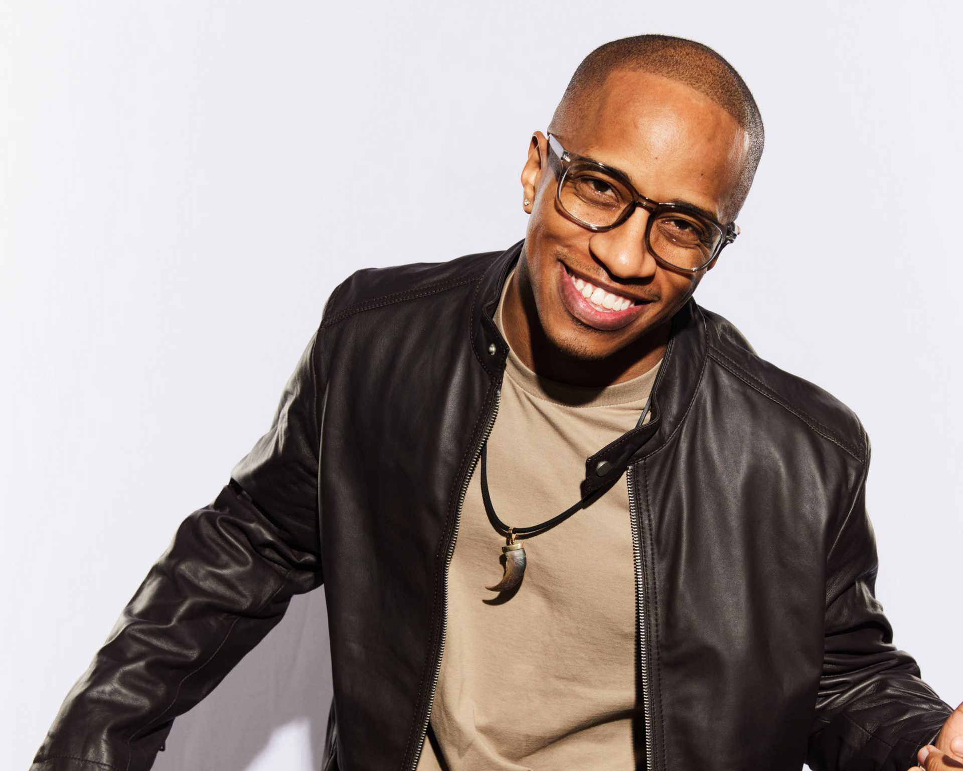 A smiling Jon Reddick in a black leather jacket and glasses with a claw-shaped pendant, set against a white background.
