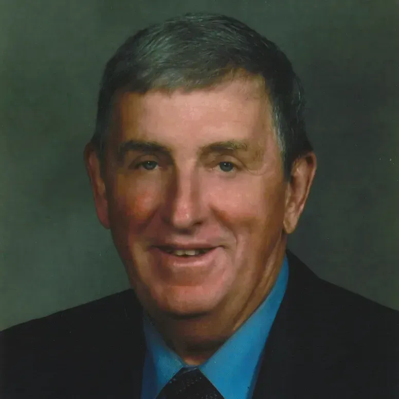 Man in a dark suit and blue shirt smiling, against a blurred background.