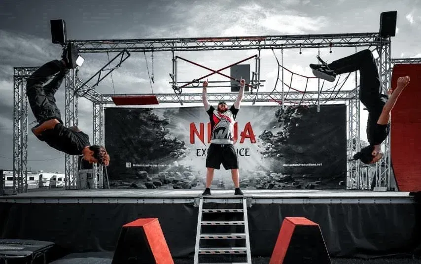 Ninja warrior obstacle course with hanging athletes and a ladder on a black stage