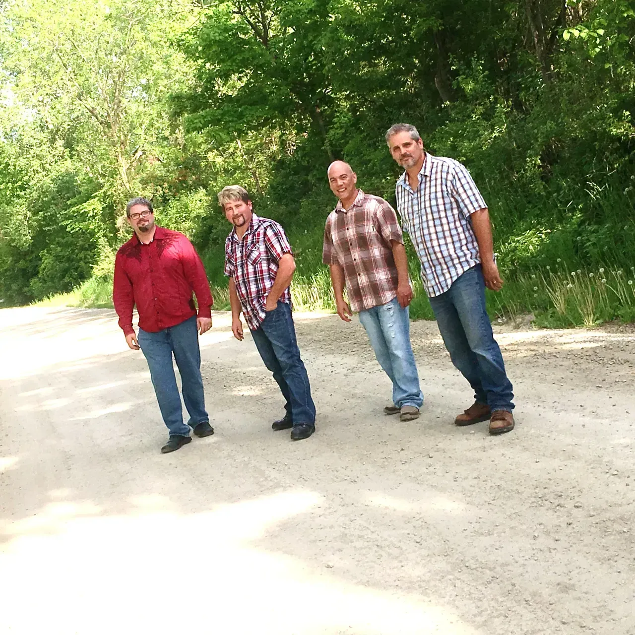 Four men stand on a dirt road surrounded by green trees, wearing casual shirts and jeans.