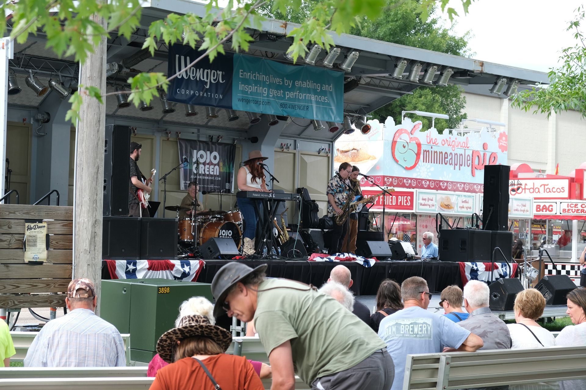 Band performing on a stage at an outdoor event, with audience seated in front.