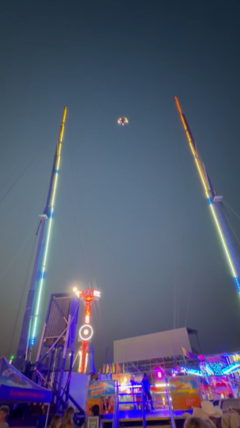 Night carnival scene with tall illuminated ride towers and a small suspended seat against a dark blue sky